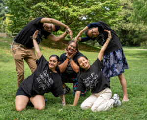 Five people making a heart formation with their bodies outside on the green grass.